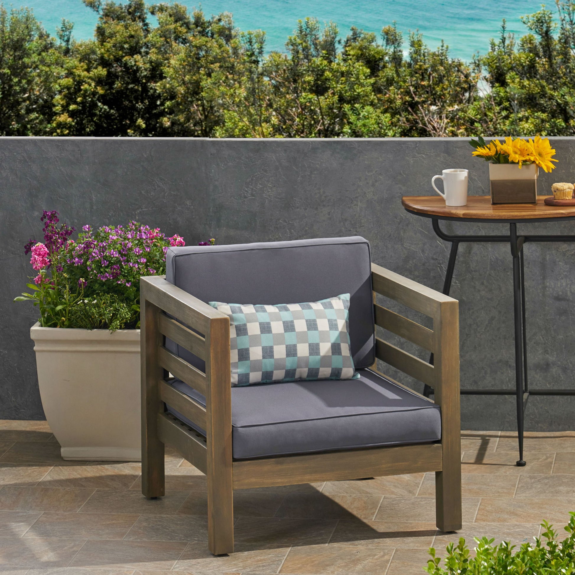 An Outdoor Acacia Wood Patio Chair with a fabric cushion sits by a table with coffee, a muffin, and flowers. Nearby is a large planter of purple flowers, all overlooking the ocean and trees.