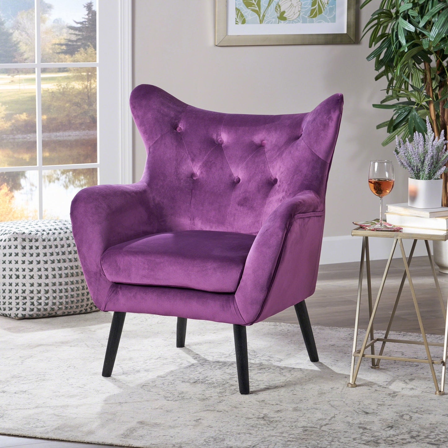 The Alyssa Mid Century Upholstered Arm Chair with wingback design in purple, featuring a tufted backrest and black legs, sits on a rug near a window. A patterned pouf, side table with wine, and framed art complete the cozy sunlit room.