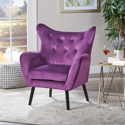 The Alyssa Mid Century Upholstered Arm Chair with wingback design in purple, featuring a tufted backrest and black legs, sits on a rug near a window. A patterned pouf, side table with wine, and framed art complete the cozy sunlit room.