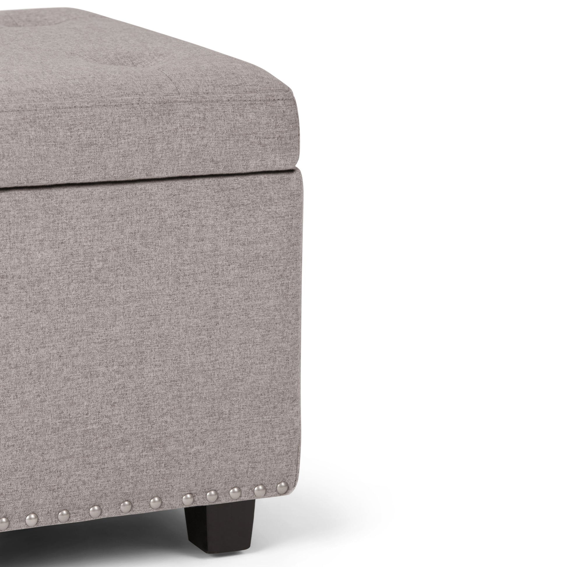 Close-up of the Hamilton Upholstered Storage Ottoman in light gray, featuring a tufted top, silver nailhead trim along the bottom edge, and short black wooden legs.