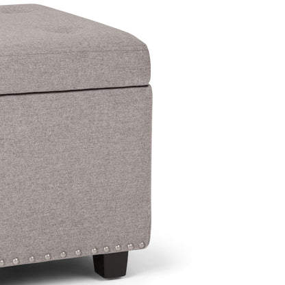Close-up of the Hamilton Upholstered Storage Ottoman in light gray, featuring a tufted top, silver nailhead trim along the bottom edge, and short black wooden legs.