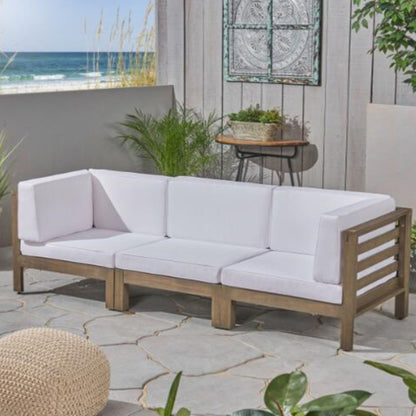 A modern patio with the Oana 3 Piece Sofa Set featuring water resistant white cushions, surrounded by green plants and a round woven pouf on stone flooring, overlooking the ocean.
