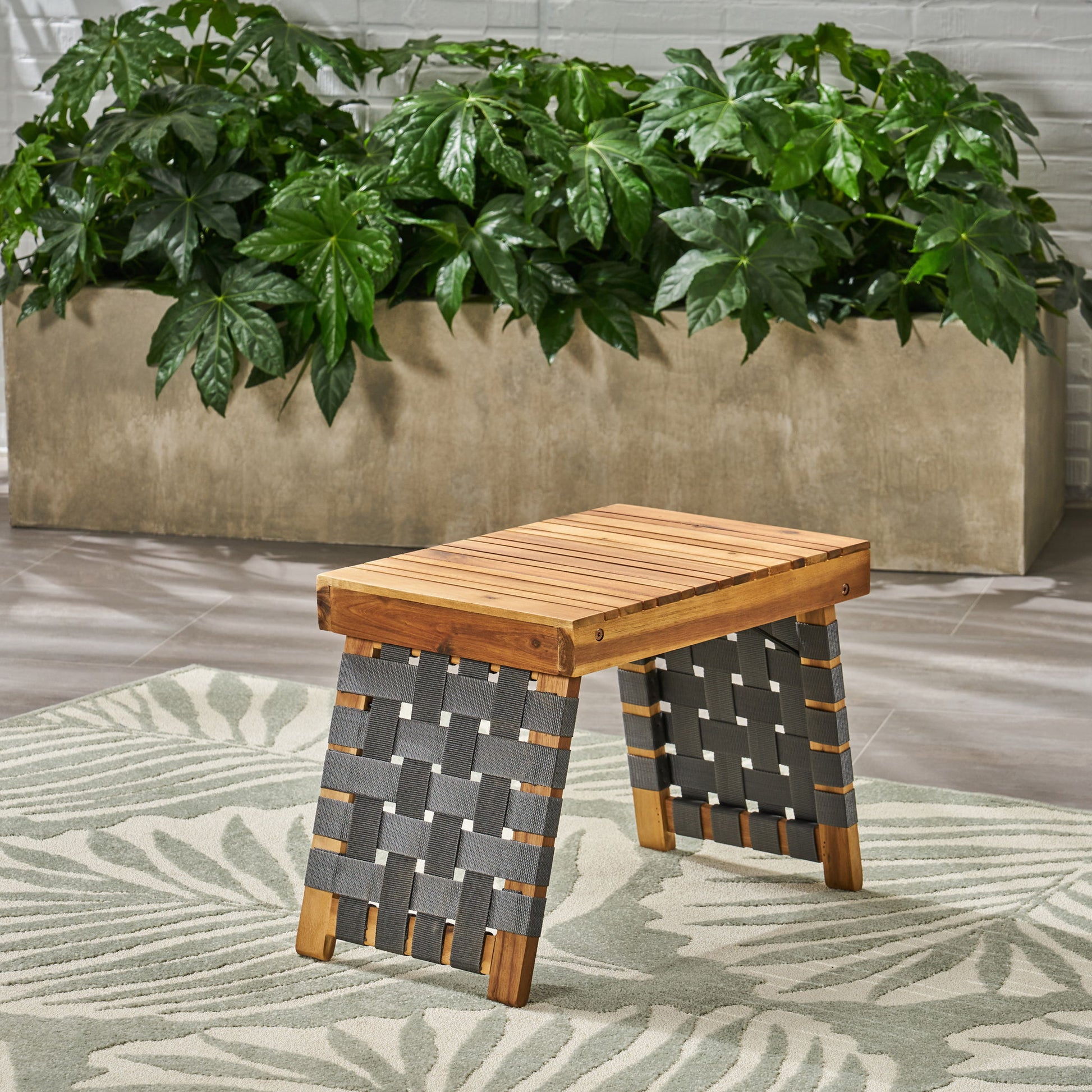 The Outdoor Wooden Foldable Side Table - Teak features a slatted seat and woven dark straps, sitting on a patterned rug with a large planter of green leafy plants in the background.