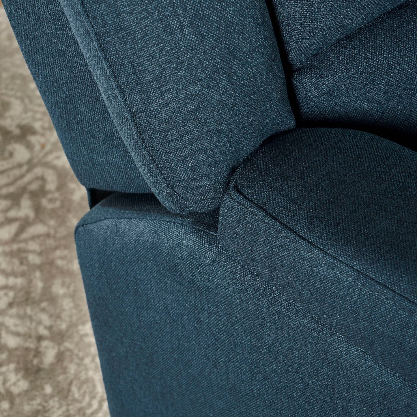 Close-up of the Contemporary Glider Recliner With Swivel Function, featuring blue fabric upholstery with visible texture on the armrest, seat, and backrest. A glimpse of a patterned beige carpet appears in the background.