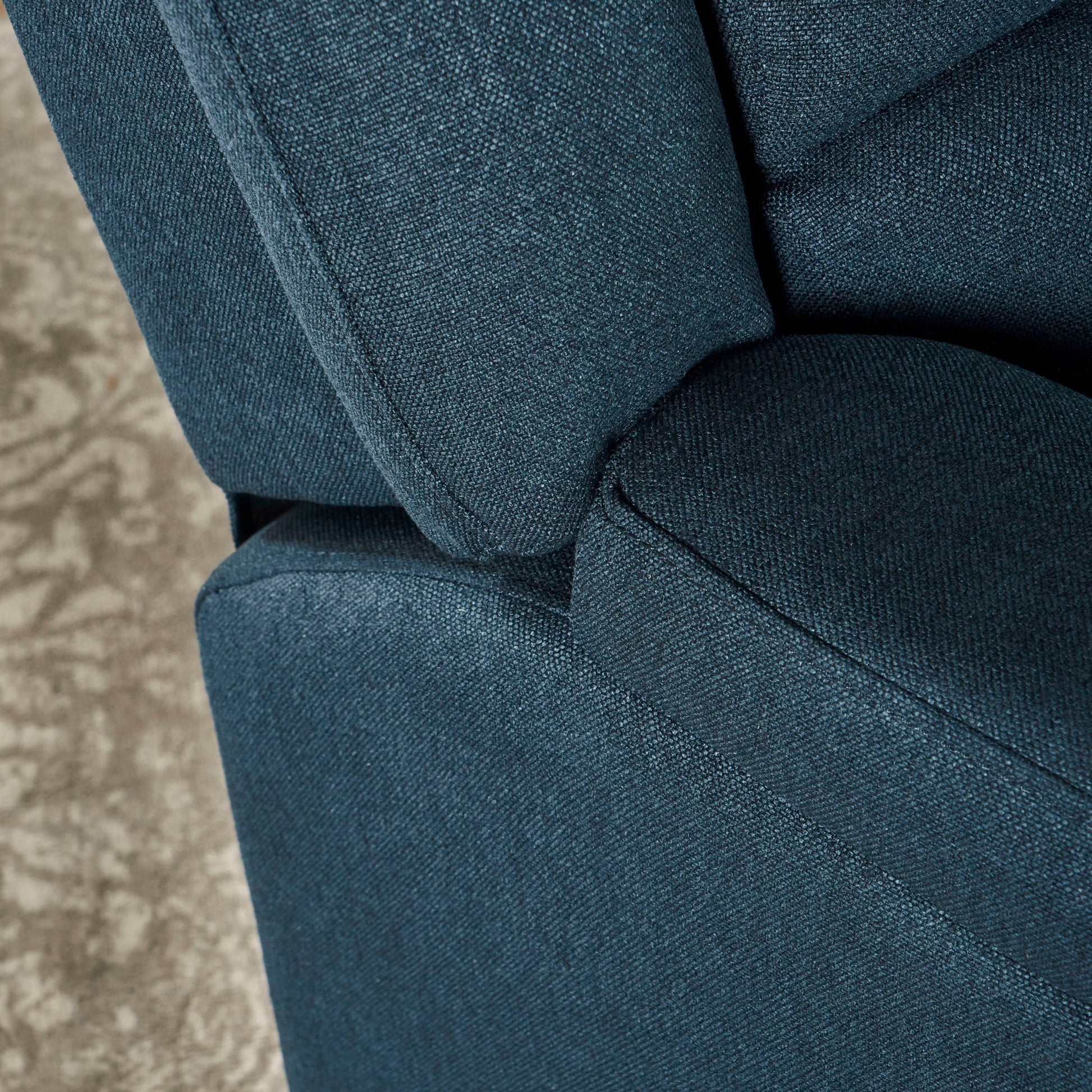 Close-up of the Contemporary Glider Recliner With Swivel Function, featuring blue fabric upholstery with visible texture on the armrest, seat, and backrest. A glimpse of a patterned beige carpet appears in the background.
