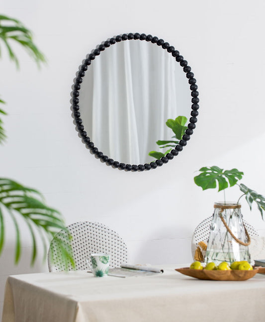 A Circle Mirror With Metal Beaded Frame in black hangs on a white wall above a dining table, set with a beige tablecloth, glass pitcher, wooden bowl of lemons, and surrounded by green plants.