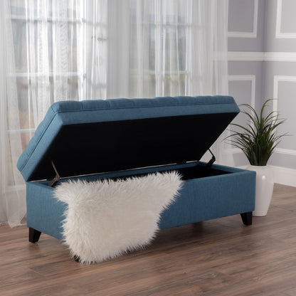 The Juliana Storage Ottoman with birch legs sits on wooden floors by a large window and potted plant. Its blue upholstery lid is open to reveal storage, while a white faux fur throw is draped over the bench.