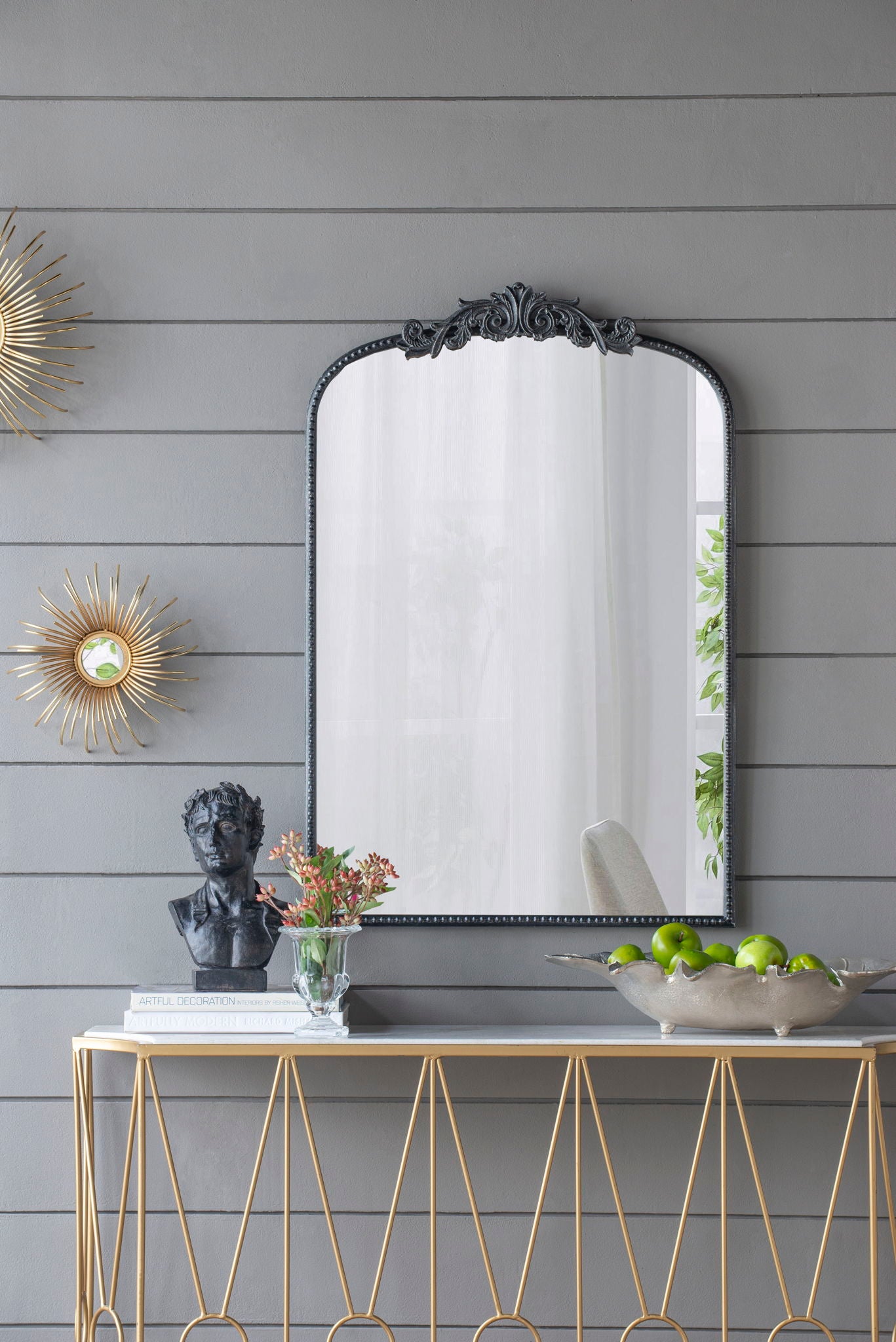 A console table with a gold geometric frame holds decor, while above it hangs the Arch Mirror, Baroque Inspired Wall Decor, flanked by sunburst decorations on a gray paneled wall.