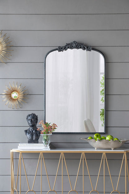 A console table with a gold geometric frame holds decor, while above it hangs the Arch Mirror, Baroque Inspired Wall Decor, flanked by sunburst decorations on a gray paneled wall.