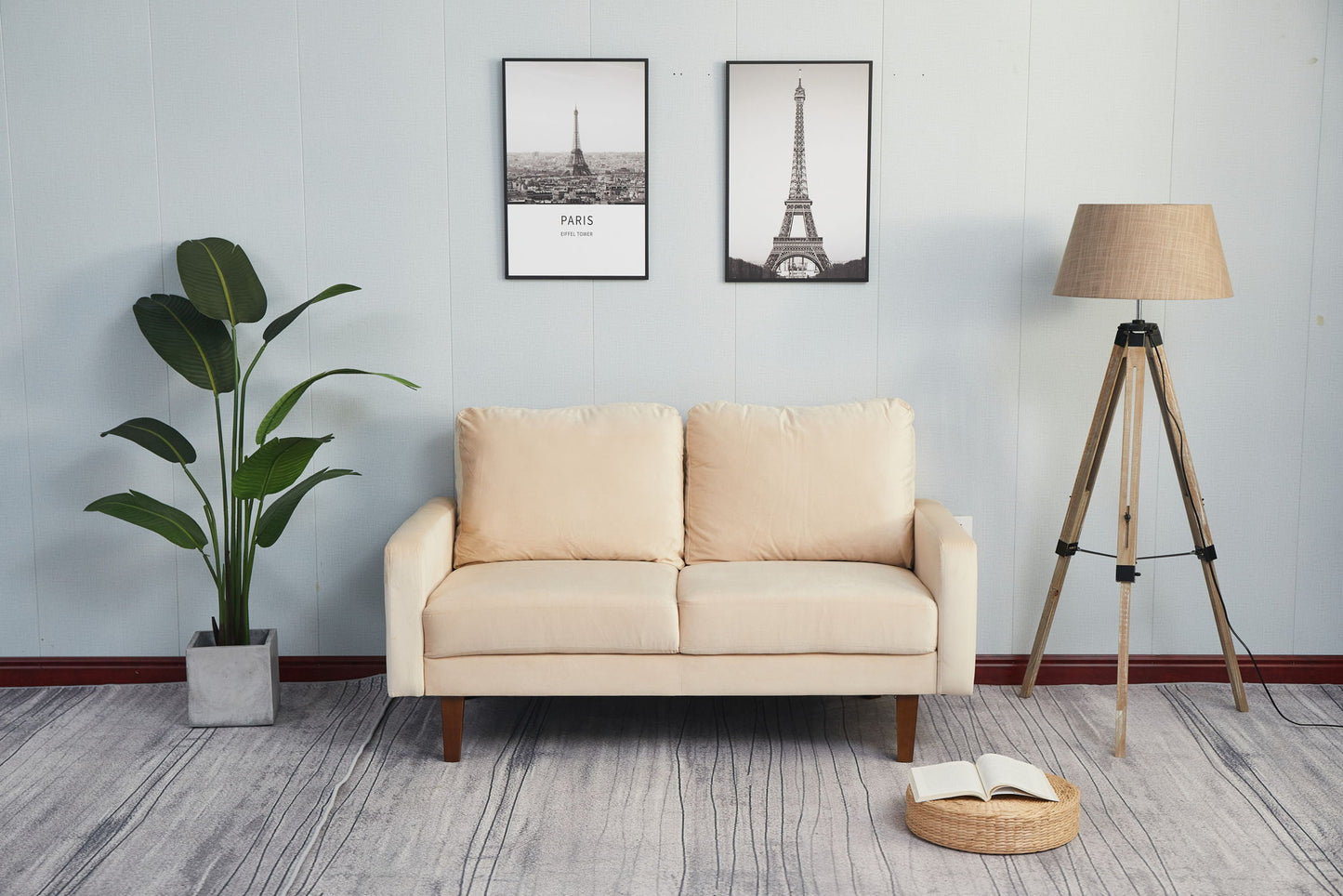 A Comfortable Loveseat With Durable Construction sits against a light blue wall with two Eiffel Tower prints above, a potted plant on the left, a floor lamp on the right, and an open book lying on the carpet in front.