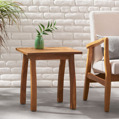 The Kinesis Outdoor Side Table with a slatted top and wood frame stands beside a cushioned chair with wooden arms, in front of a white brick wall and next to a leafy plant, topped with a green vase holding leafy branches.