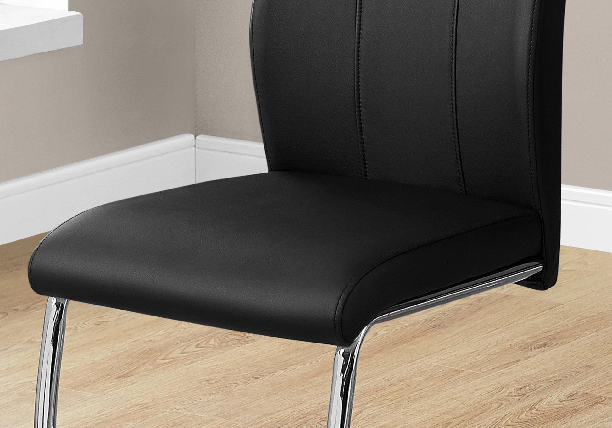 A set of 2 contemporary black dining chairs with cushioned seats and backrests, chrome metal legs, shown on a light wood floor near a beige wall and window ledge.