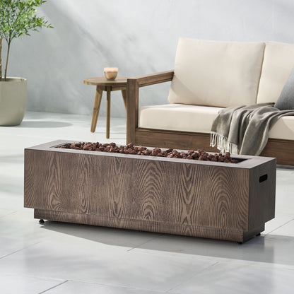 The Wellington Square Iron Fire Pit With Wood Pattern Design and 50,000 Btu output sits on a tiled patio with lava rocks on top. Behind it are a beige outdoor sofa with a gray throw, a small wooden table, potted plant, and candle.