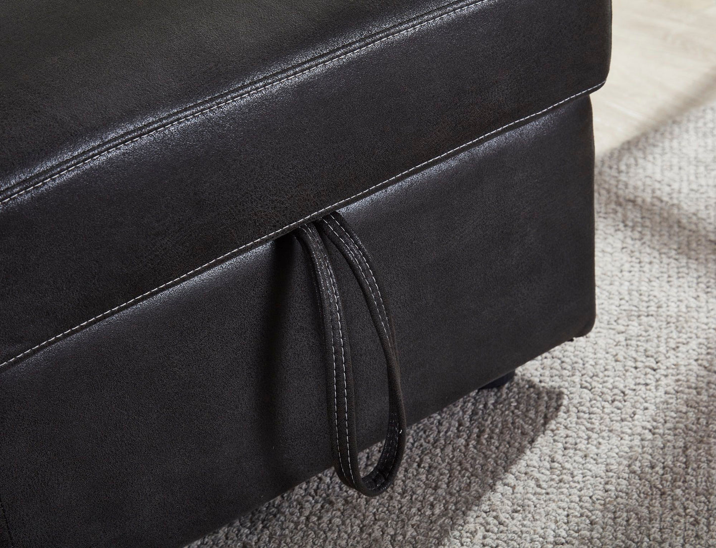 A close-up of the Comfort Sectional Sofa Bed, featuring dark upholstery and a stitched handle detail, shown atop a light-colored textured carpet.