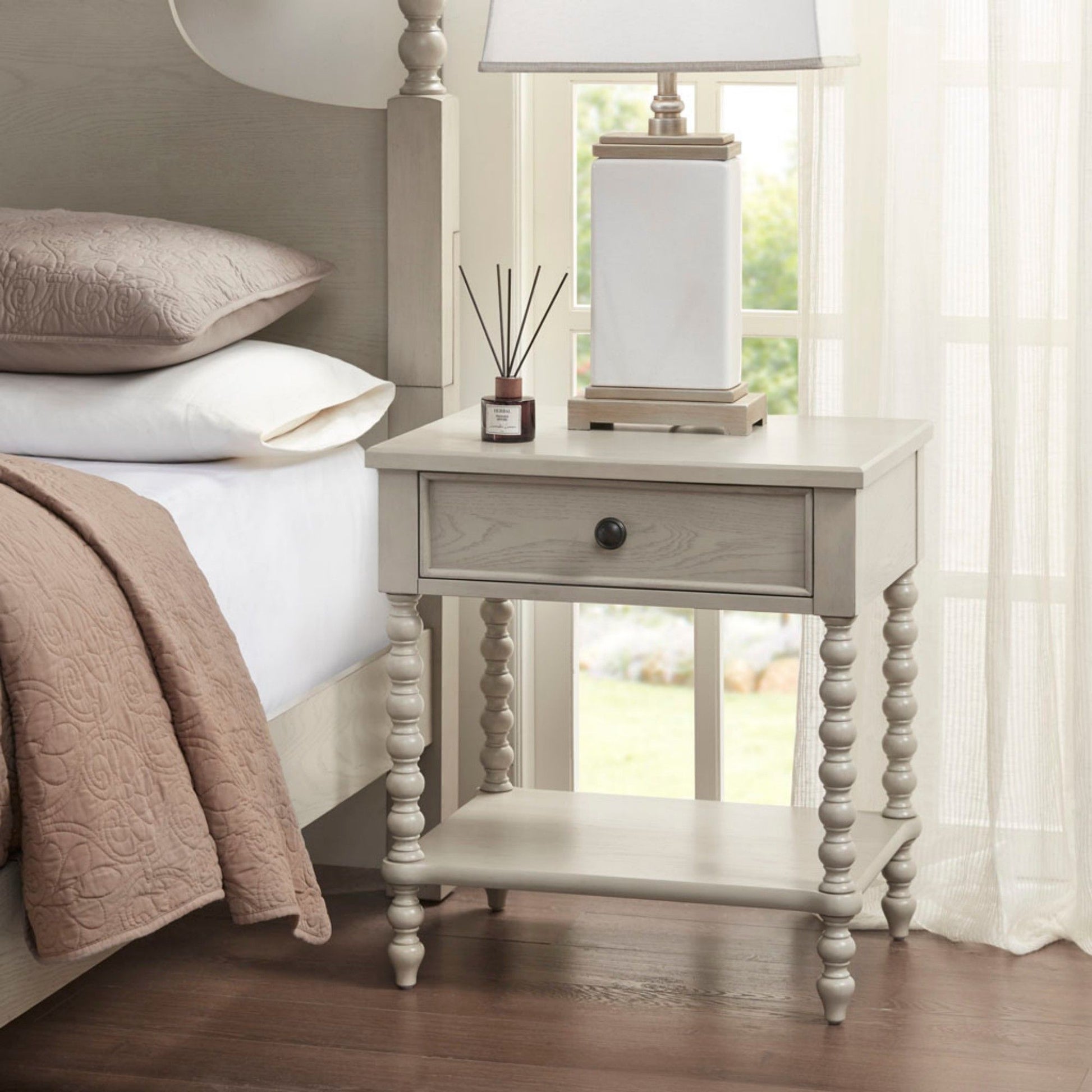 A Traditional Durable Nightstand Table With Drawer And Shelf with turned legs is placed beside a bed with tan bedding. On top are a reed diffuser and a white lamp, with sunlight streaming through sheer curtains behind the scene.