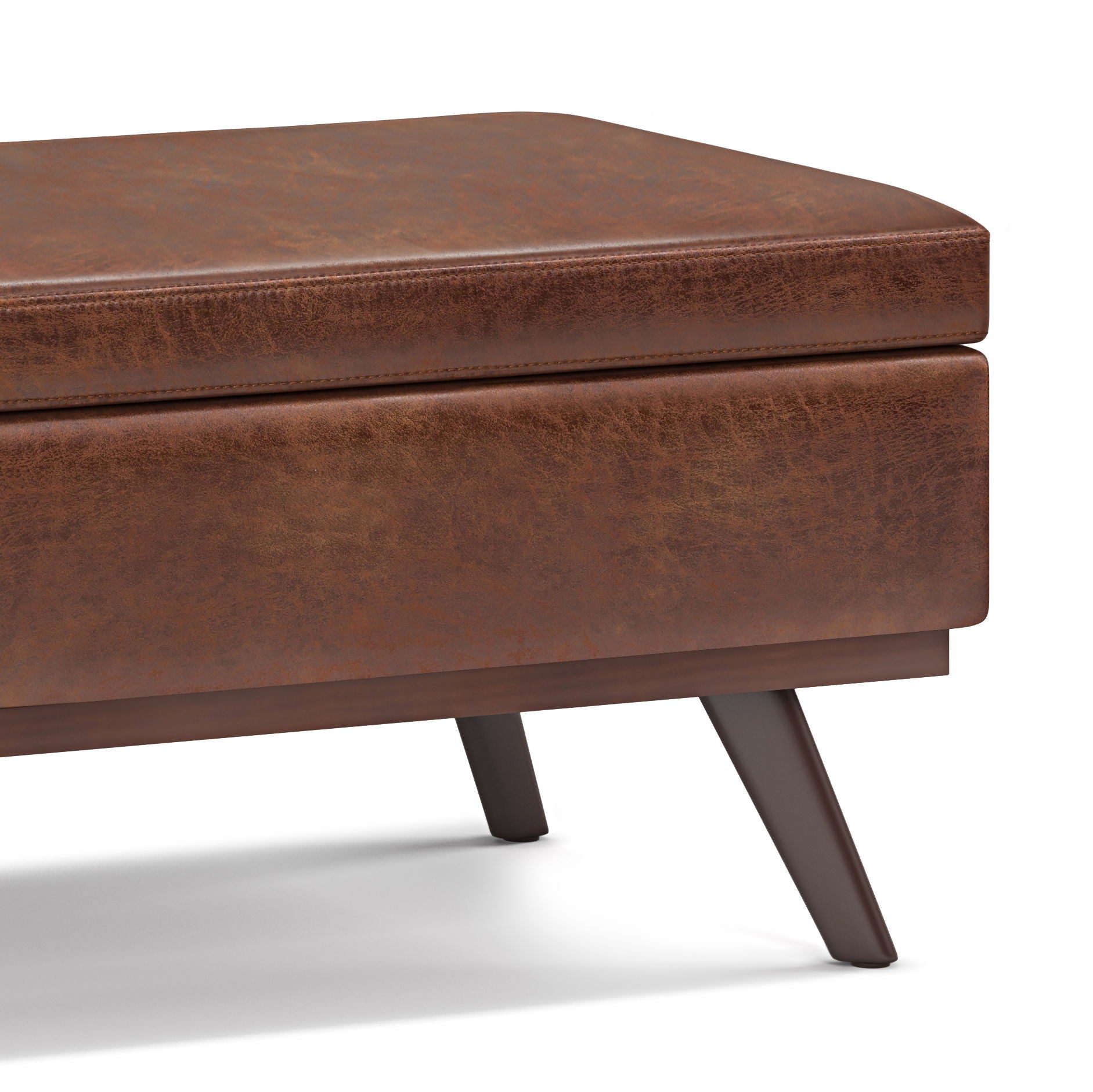 Close-up of the Owen Lift Top Large Coffee Table Storage Ottoman in brown distressed leather with angled wooden legs and a visible seat corner, showcasing its mid-century style against a white background.