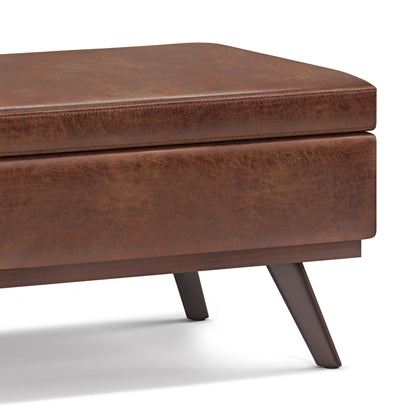 Close-up of the Owen Lift Top Large Coffee Table Storage Ottoman in brown distressed leather with angled wooden legs and a visible seat corner, showcasing its mid-century style against a white background.