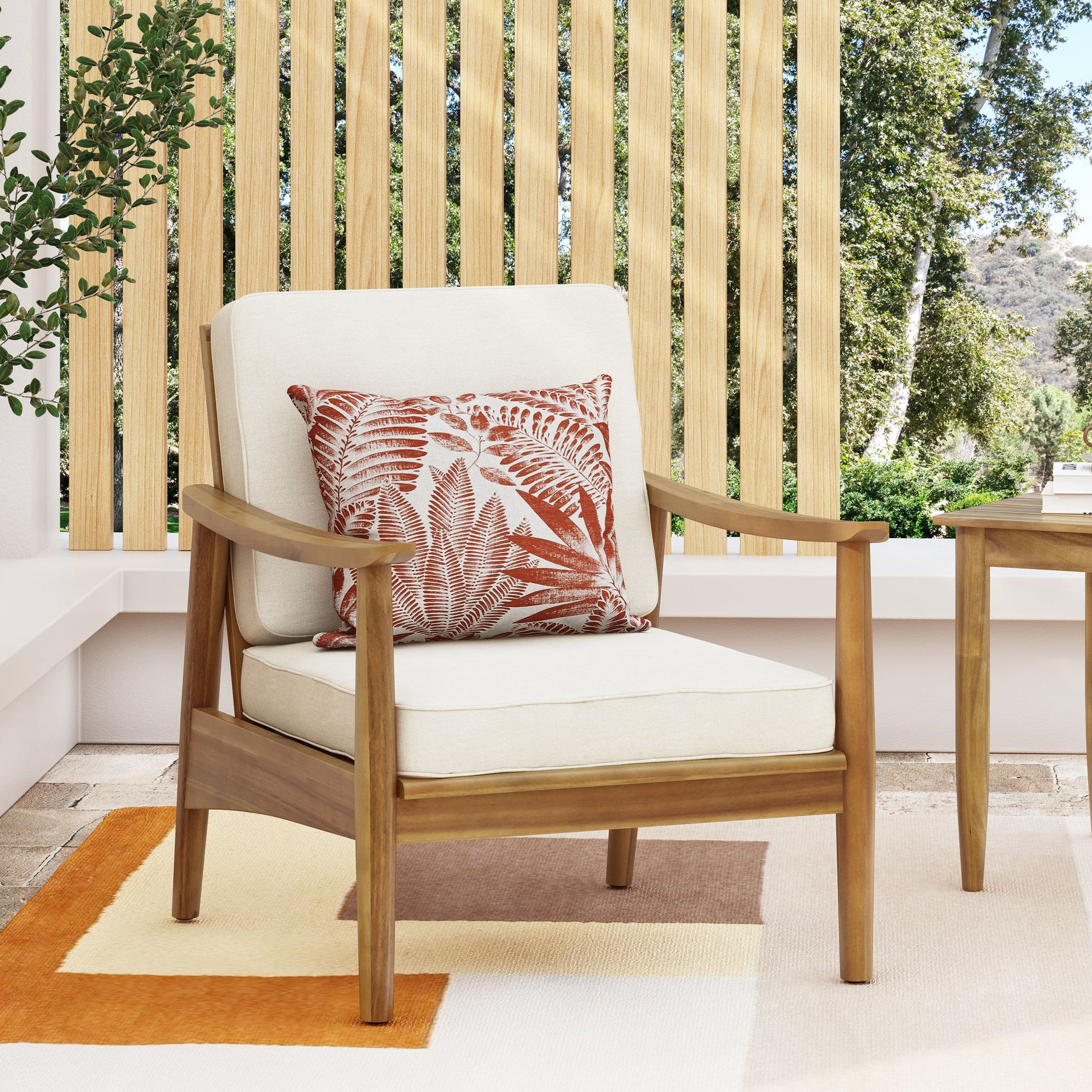 The Emmry Outdoor Acacia Wood Club Chair, featuring cream cushions and a rust-fern patterned pillow, sits on a patio with vertical wooden slats and lush green trees in the background.