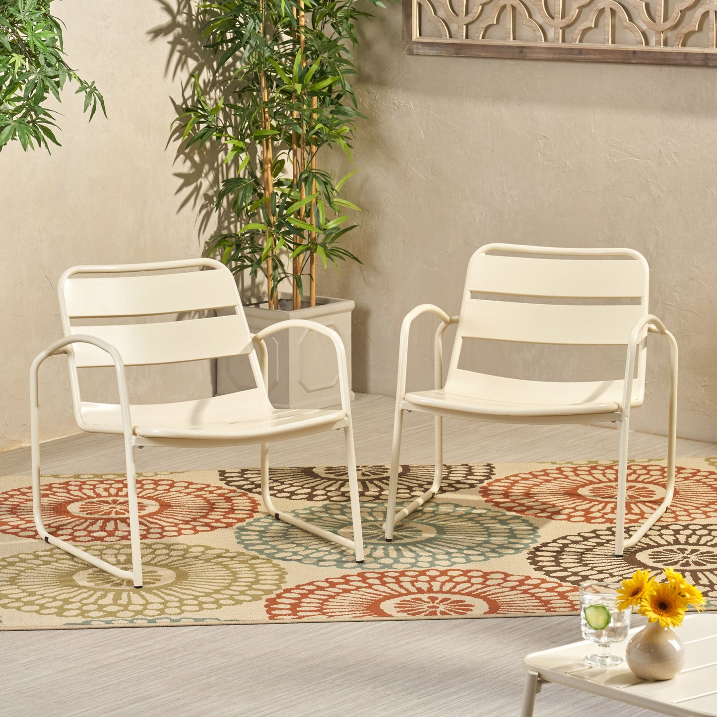 Two Conrad - Chair Outdoor Seating pieces in white sit on a patterned floral rug. Nearby are potted plants, a small table with sunflowers, and drinks in a bright, modern indoor space.
