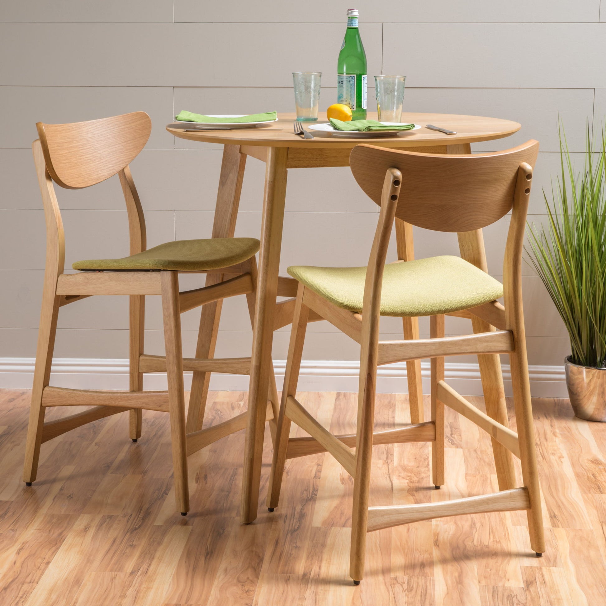 A small round wood table with two Retro Modern Counter Height Chairs featuring light green cushions. The table holds plates, glasses, a bottle of sparkling water, and utensils. A potted plant is nearby on the wooden floor.