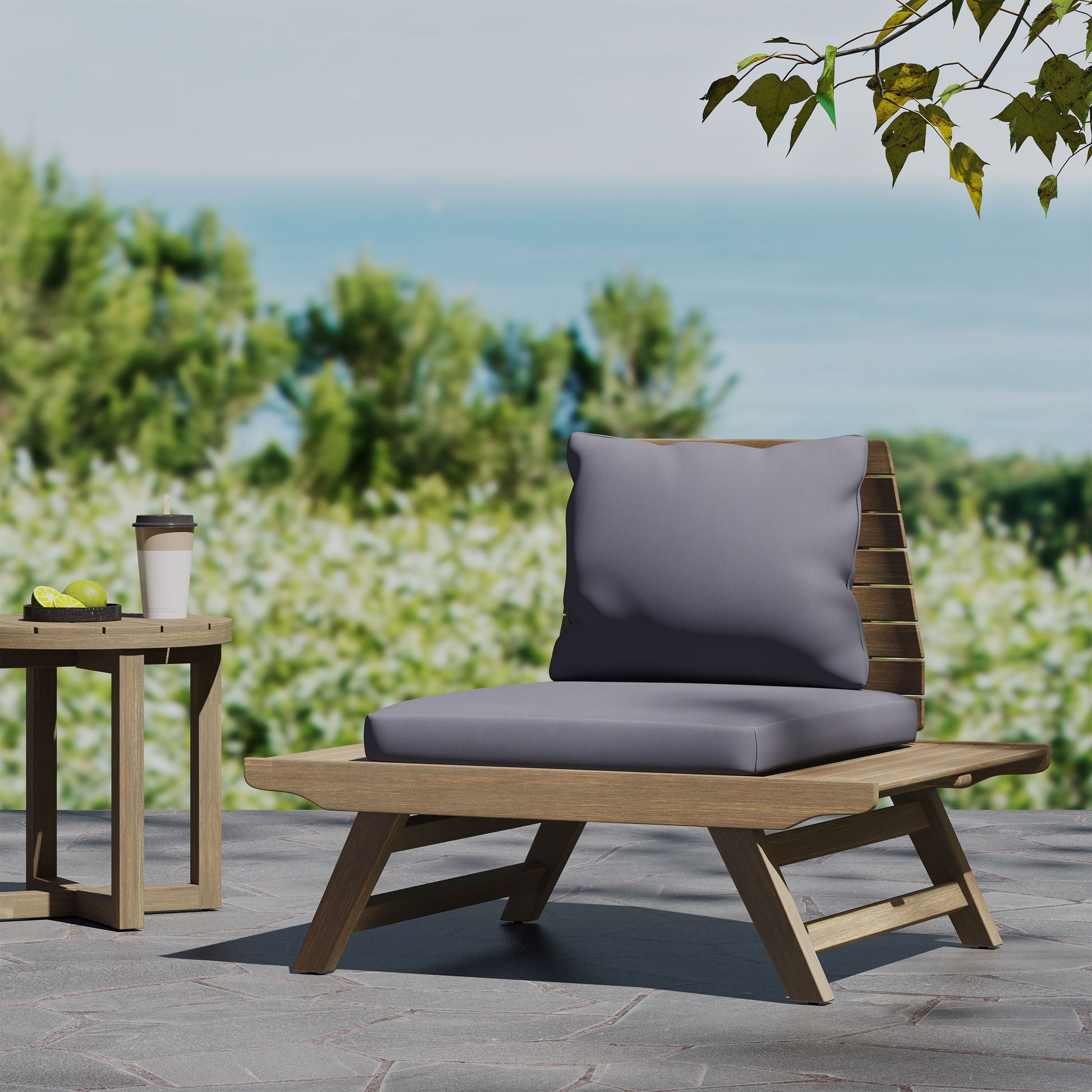 The Sedona Outdoor Club Chair with a slatted design and gray cushions rests on a stone patio beside a small round table holding a takeaway coffee cup and green fruit, with lush greenery and water in the background.