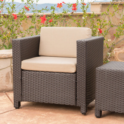 A Stylish Outdoor Club Chair With Weather Resistant Cushions in brown wicker and beige cushions sits on a stone patio, with red hibiscus flowers, a stone wall, and distant hills in the background.