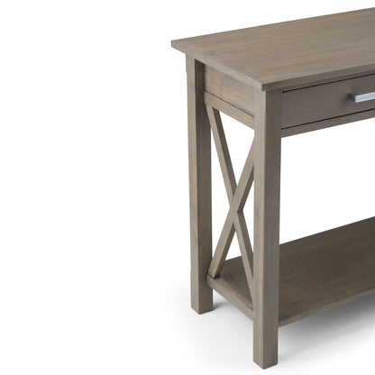 The Kitchener - Handcrafted Table is a wooden console with a gray-brown finish, X-shaped side detail, silver-handled drawer, and lower shelf. Only the side and part of the front are visible against a white background.