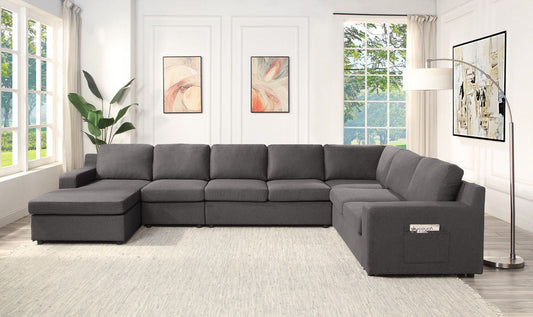 A modern living room features the Waylon - Linen Sectional Sofa in dark gray, a light rug, floor lamp, potted plant, and two abstract paintings on white walls. Large windows fill the space with natural light and showcase views of greenery.