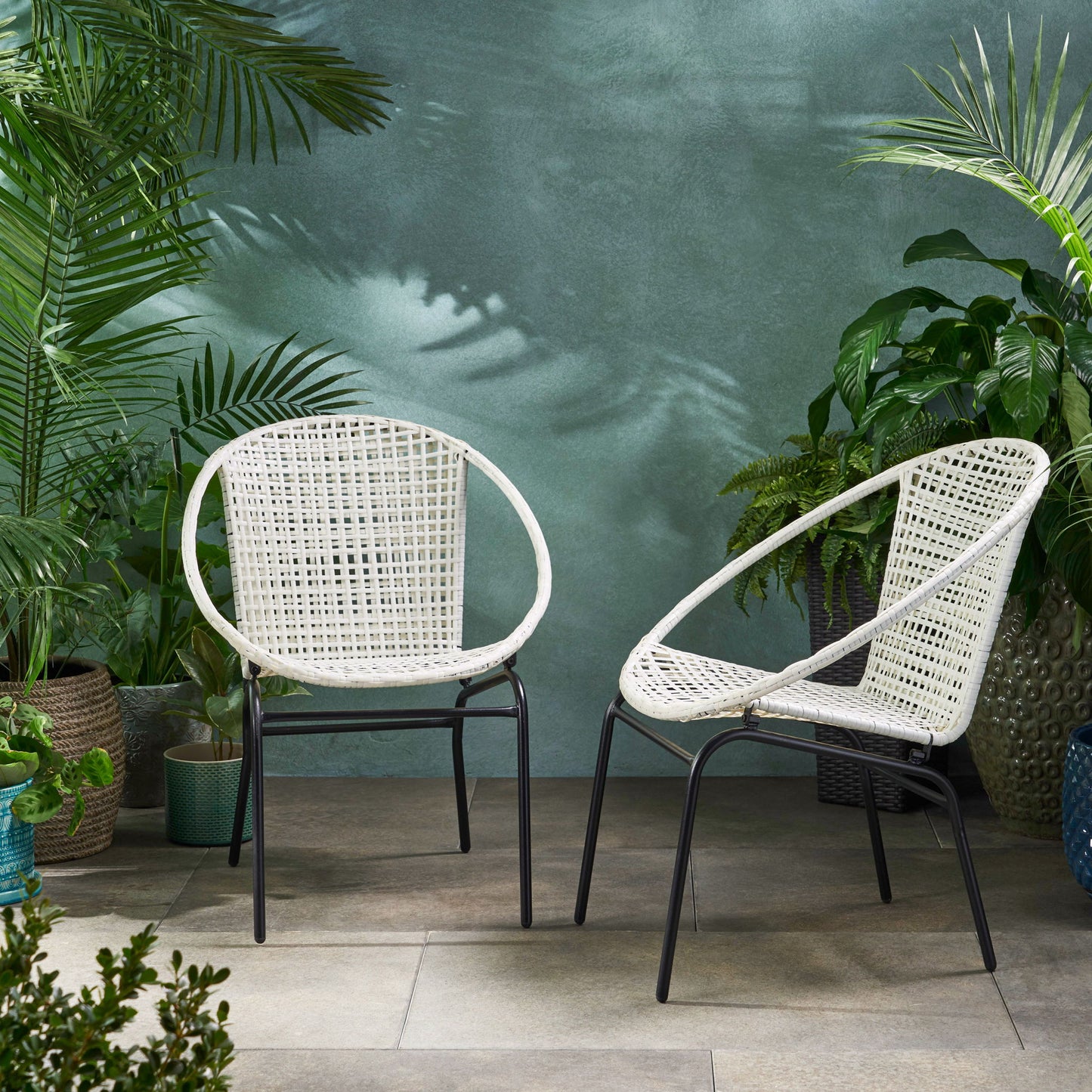 Two Java - Chair With Comfortable Design in white wicker with black metal legs are on a tiled patio, surrounded by lush green potted plants against a textured green backdrop.
