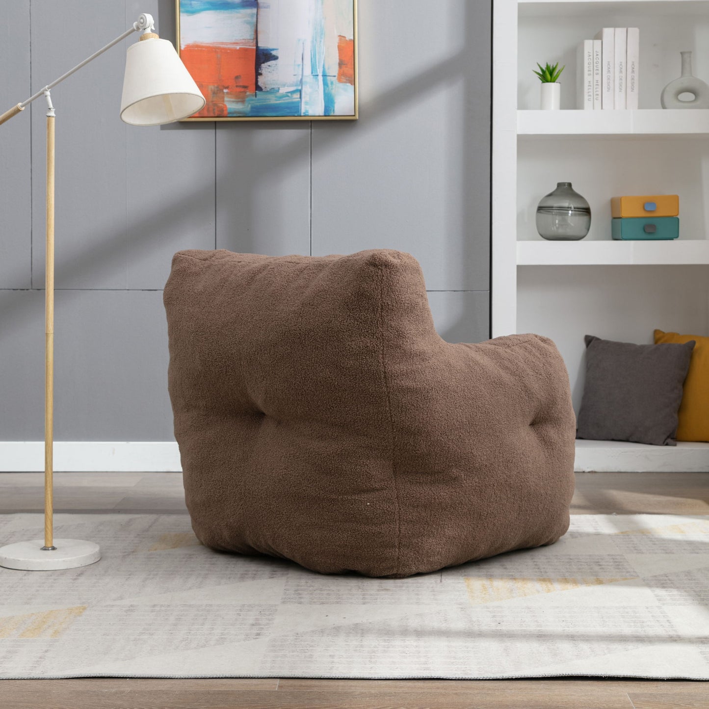A Soft Teddy Fabric Tufted Foam Bean Bag Chair with teddy fabric sits on a light area rug in a modern living room, complemented by a floor lamp, abstract wall art, and white shelves holding books and decor.