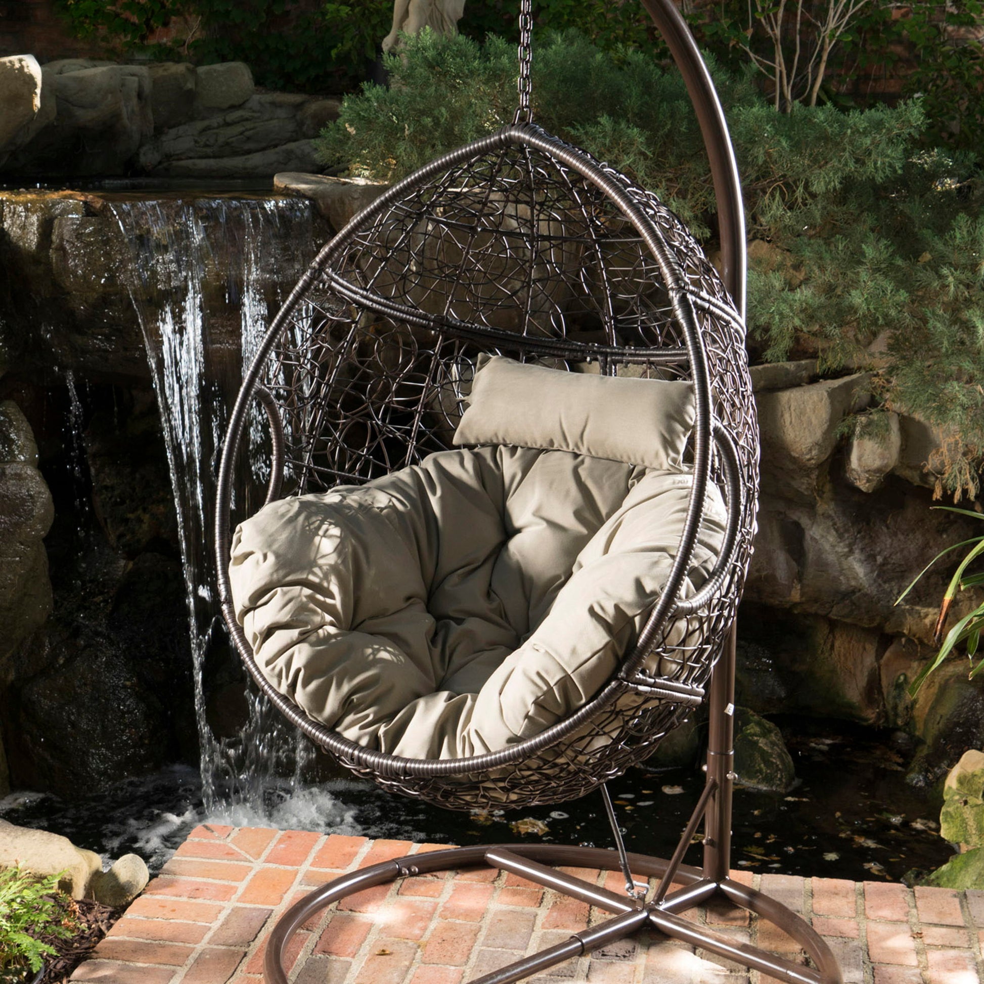 The Kylie Boho Style Hanging Basket Chair Rattan with water-resistant cushions is placed on a brick patio near a small waterfall surrounded by greenery.