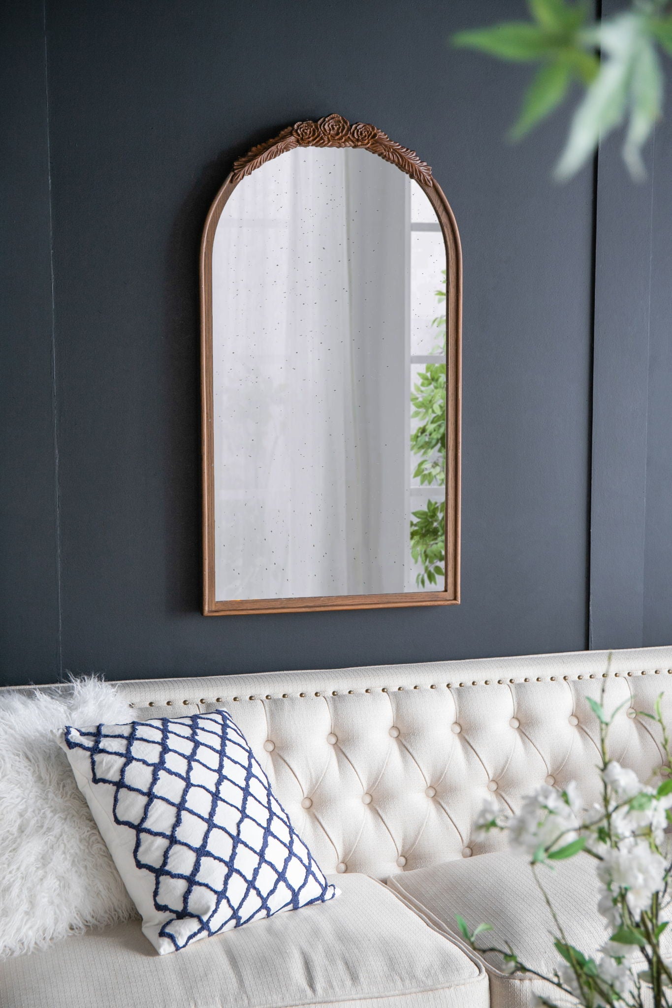 A Hand Carved Rose Antique Mirror Frame, Wood Arch Mirror Wall Decor for Living Room, hangs on a dark wall above a cream tufted sofa with a white and blue pillow; green leaves appear in the foreground.