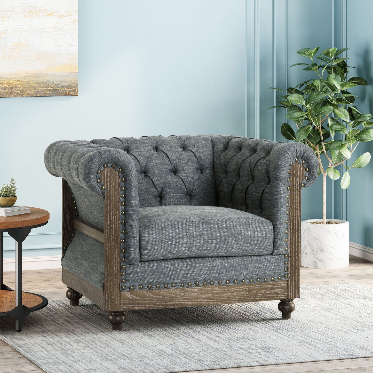 The Fabric Upholstered Club Chair With Scrolled Tuxedo Arms, featuring tufted gray fabric, wood trim, and nailhead accents, sits on a light rug next to a round wooden side table and potted plant against a light blue wall.