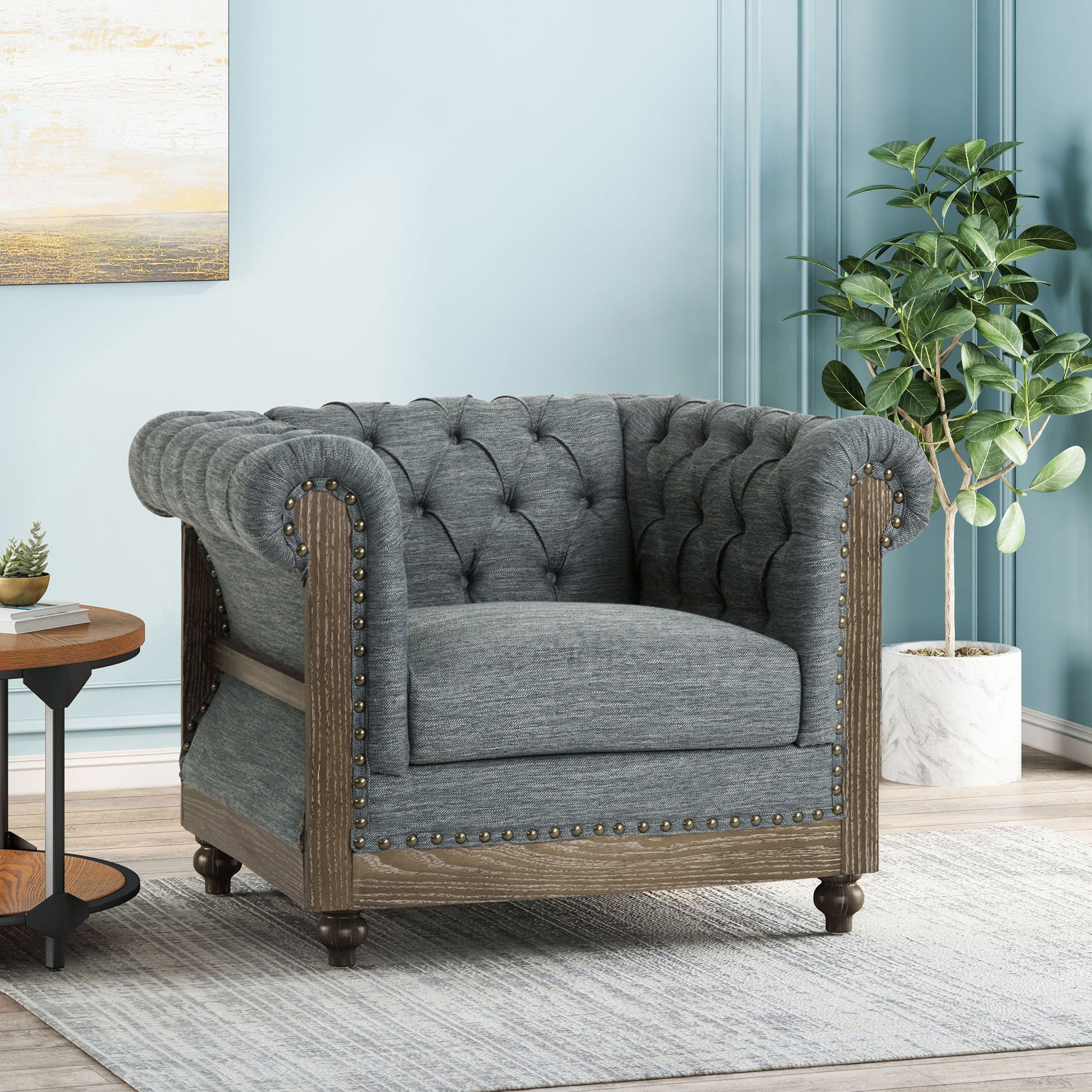 The Fabric Upholstered Club Chair With Scrolled Tuxedo Arms, featuring tufted gray fabric, wood trim, and nailhead accents, sits on a light rug next to a round wooden side table and potted plant against a light blue wall.