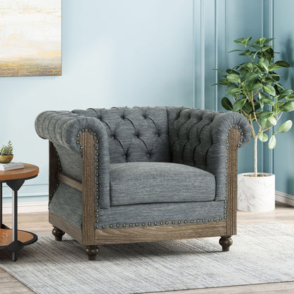 The Fabric Upholstered Club Chair With Scrolled Tuxedo Arms, featuring tufted gray fabric, wood trim, and nailhead accents, sits on a light rug next to a round wooden side table and potted plant against a light blue wall.