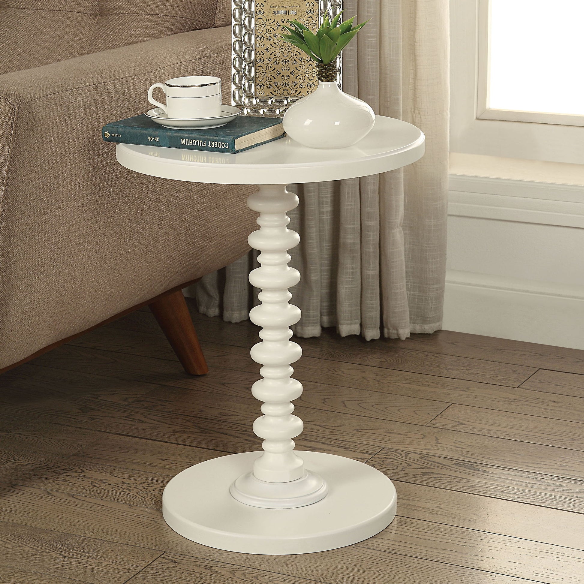 The Acton Accent Table in white, with a sculpted pedestal base, holds a white vase with greenery, books, and a cup. It sits next to a beige sofa on a wooden floor near a window.