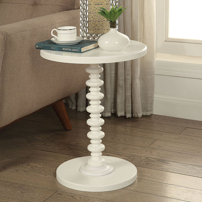 The Acton Accent Table in white, with a sculpted pedestal base, holds a white vase with greenery, books, and a cup. It sits next to a beige sofa on a wooden floor near a window.