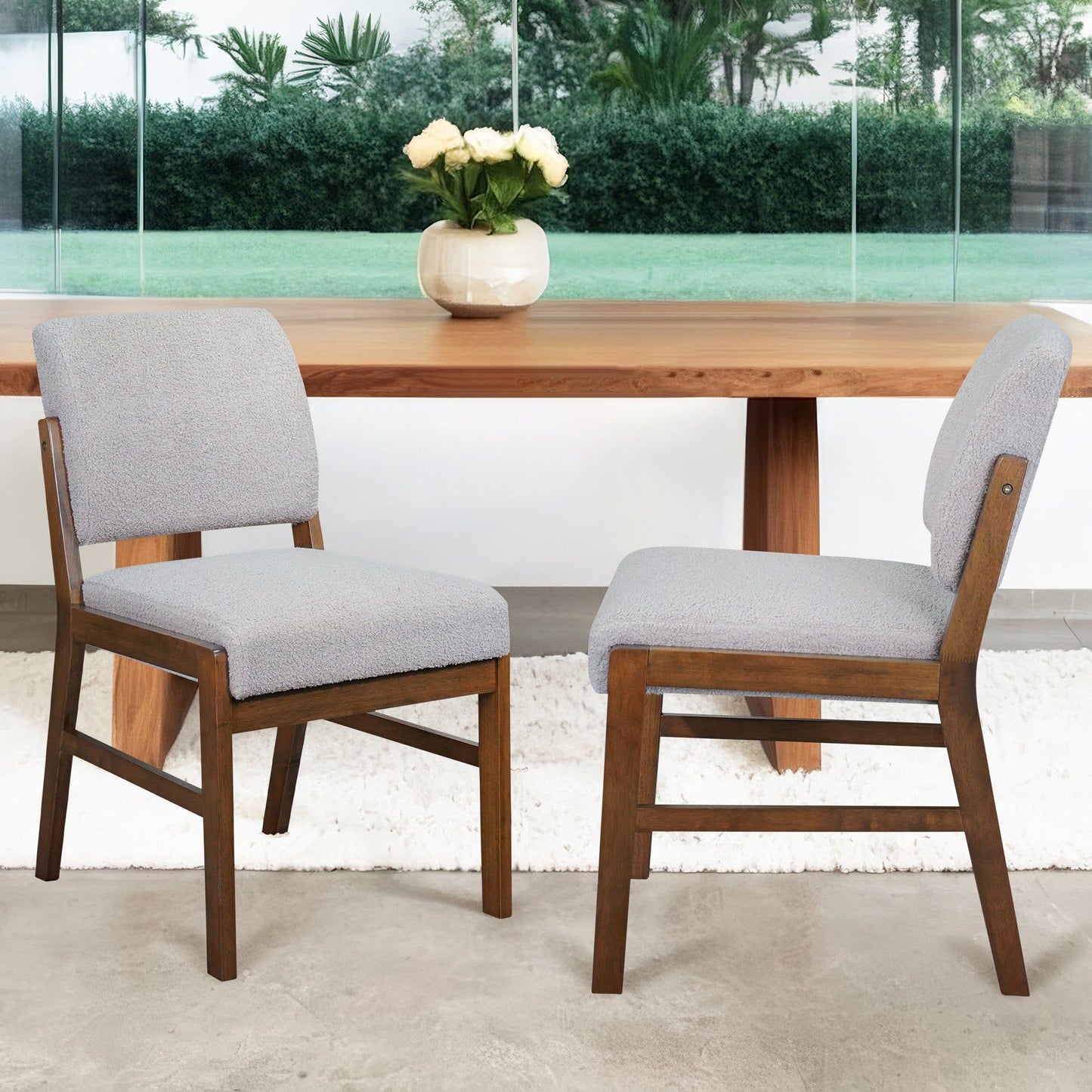 Two Caldwell Dining Chairs (Set of 2) in Light Gray are placed in front of a wooden dining table with a vase of white flowers, indoors near a large window overlooking greenery.