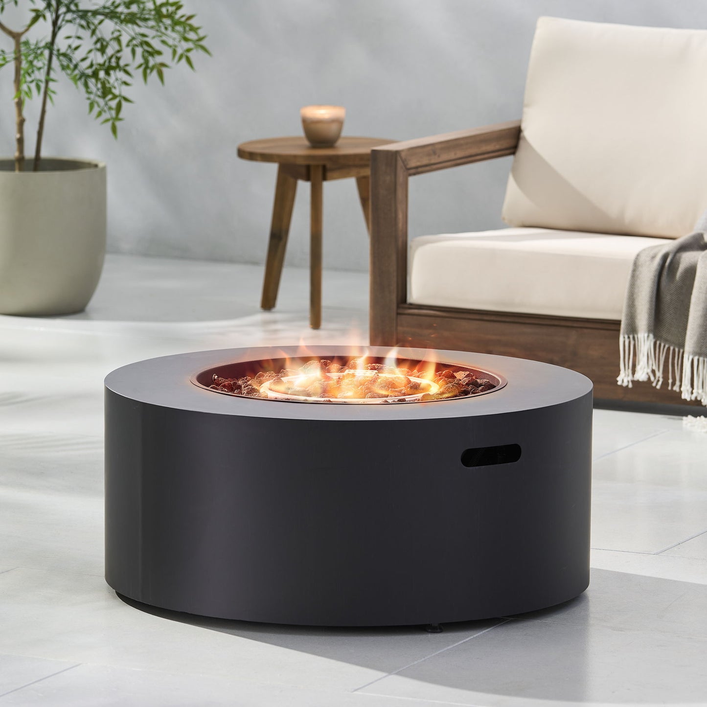 The Round Iron Fire Pit With 40,000 Btu, finished in sleek black, burns with visible flames on a light patio next to a wooden chair with cream cushions, a small side table, and a potted plant in the background.