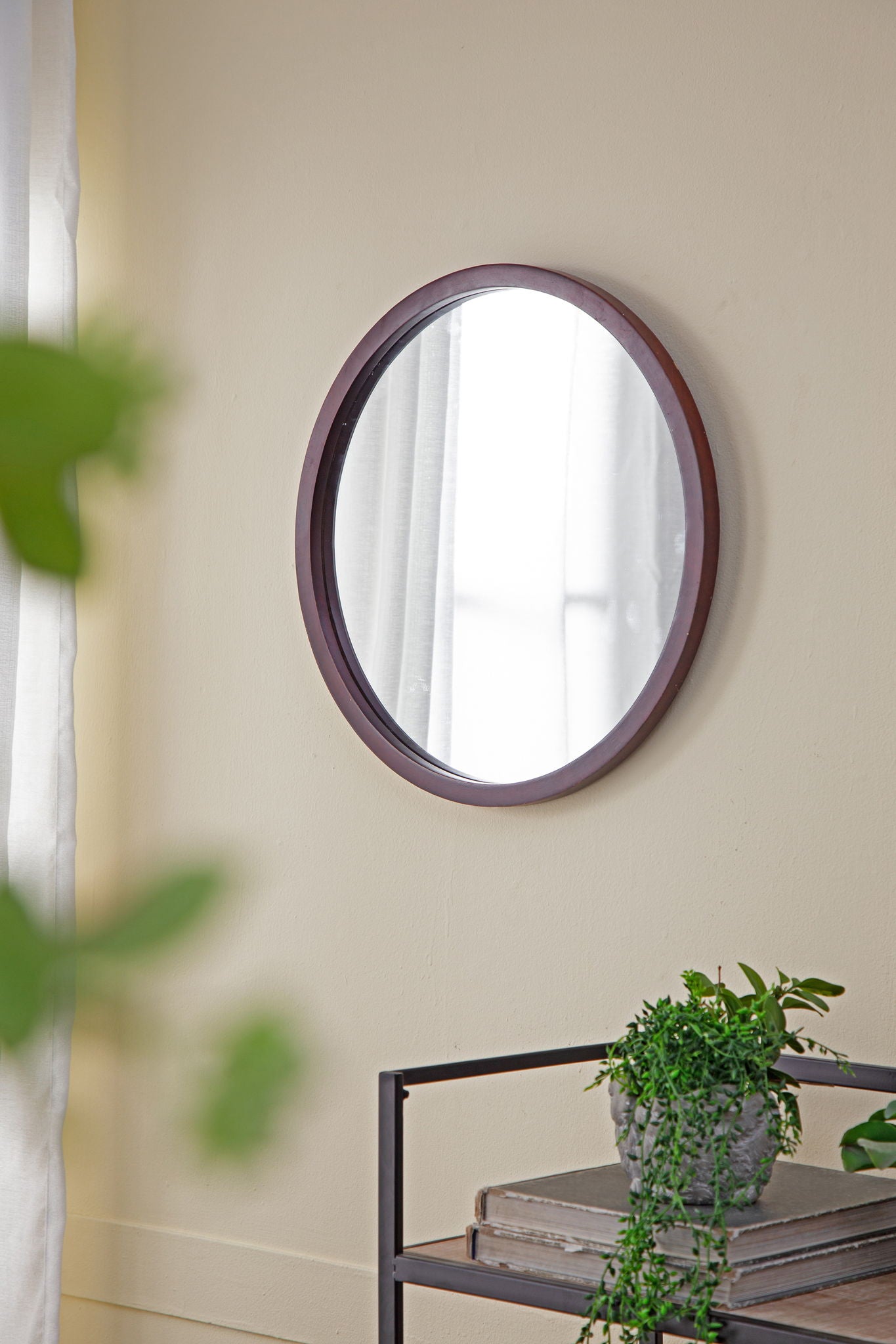 A Circle Wall Mirror With Wooden Frame hangs on a beige wall. Below it, a metal shelf displays a gray pot with trailing plants and books. A white curtain and green foliage are partly visible nearby in the living room.