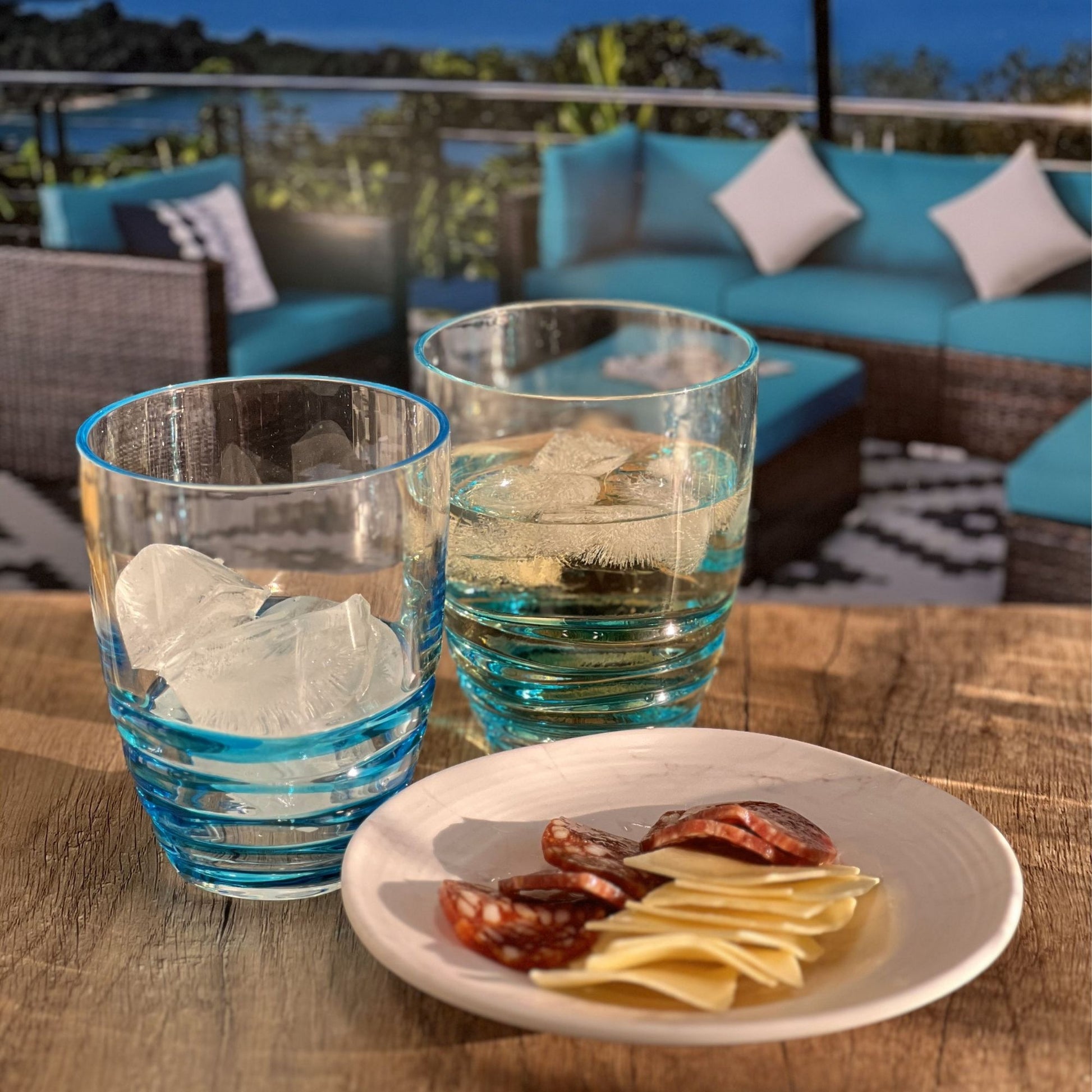 Two Leading Ware Swirl Acrylic Drinking Glasses with ice and water sit on a wooden table beside a small plate with cheese and salami. Outdoor patio seating with turquoise cushions appears in the background.