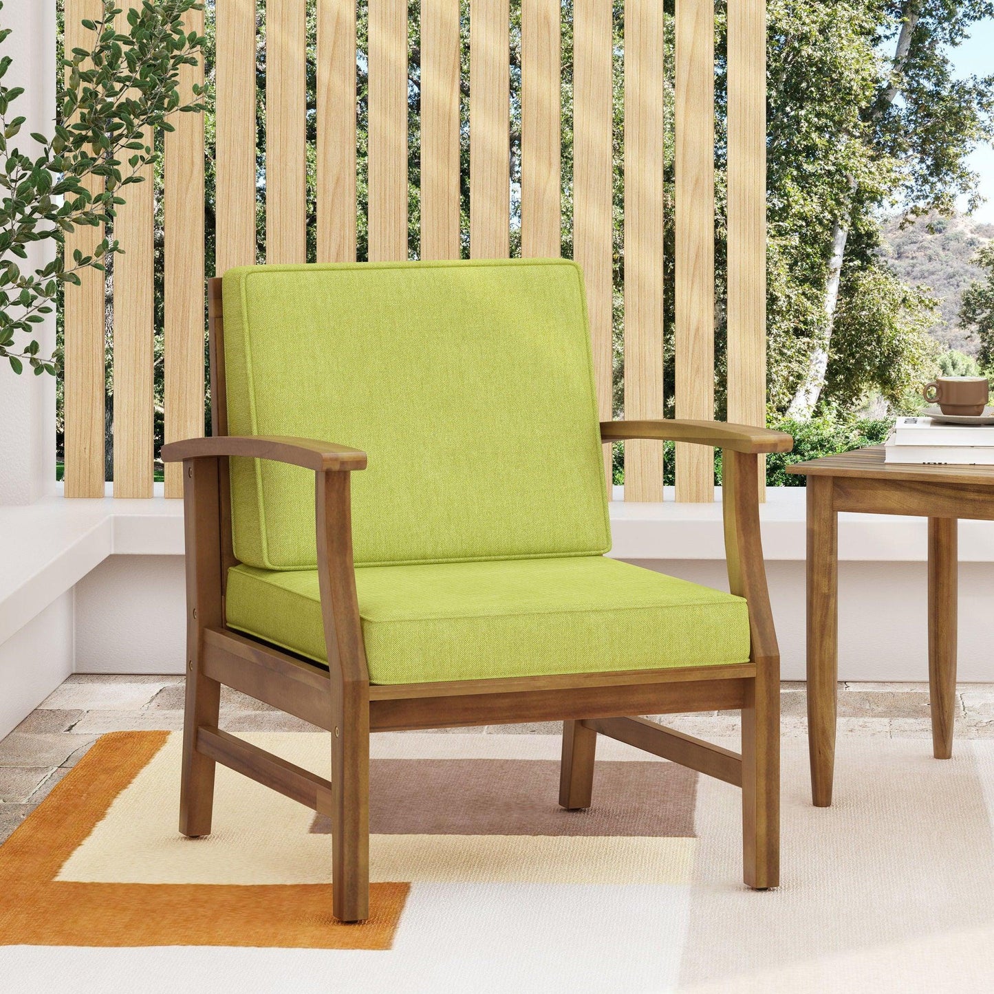 The Perla - Contemporary Club Chair with water-resistant cushions sits on a patio rug beside a wooden table. Vertical wooden slats and leafy trees in the background create a cozy outdoor setting.
