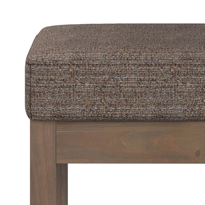 A close-up of the Milltown Upholstered Ottoman Bench highlights its cushioned, textured brown seat and square wooden legs, showing the top and part of the frame.