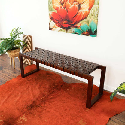 The Christina Leather Bench in Cognac and Dark Brown sits on an orange rug, accented by a potted plant and bold red floral wall art in a bright, stylish room with wood floors.