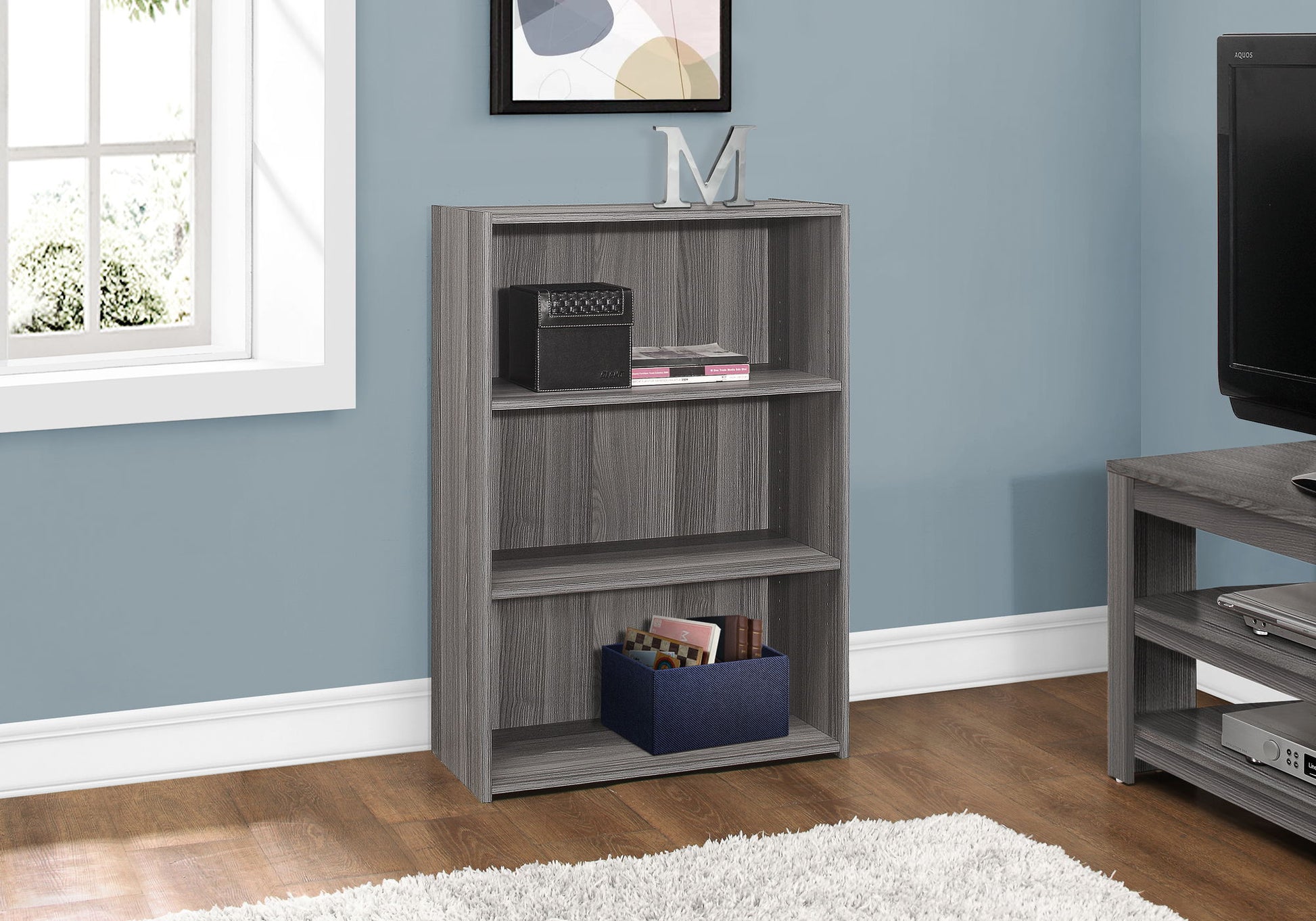 A gray 4-tier transitional bookshelf for office is placed against a blue wall. On the shelves are a black box, books, and a blue storage bin, with a decorative "M" on top. A TV and window are close by.