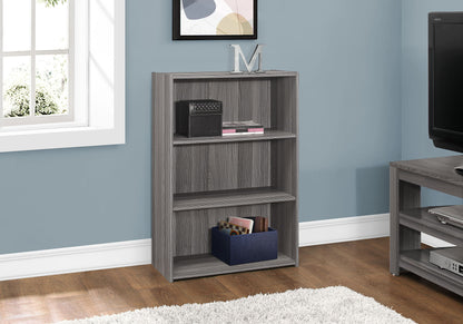 A gray 4-tier transitional bookshelf for office is placed against a blue wall. On the shelves are a black box, books, and a blue storage bin, with a decorative "M" on top. A TV and window are close by.