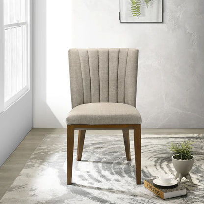 Two Elmwood Stylish Dining Chairs with beige upholstery and wooden legs stand on a patterned rug in a bright, modern room. Nearby is a small table with a plant, cups, and a book; a framed botanical print adorns the wall.