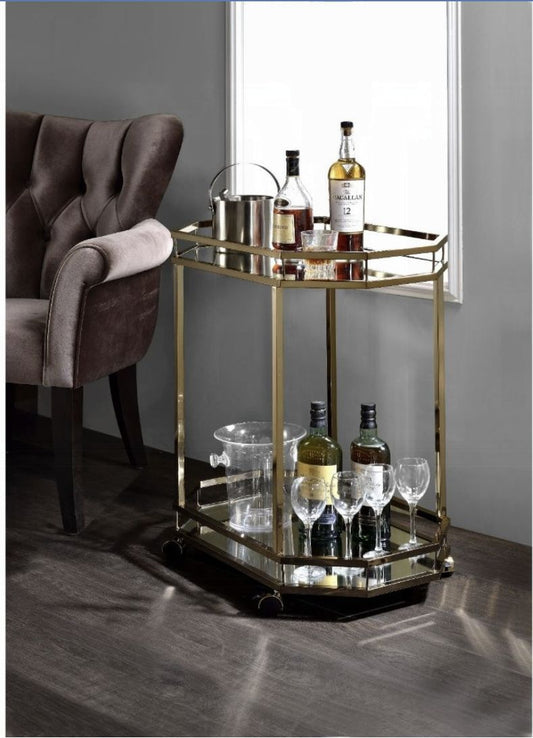 The Lacole Mirrior Serving Cart in Champagne, featuring glass shelves and a gold frame, displays liquor bottles, glassware, and an ice bucket beside a plush dark brown armchair on a dark wood floor against a gray wall.