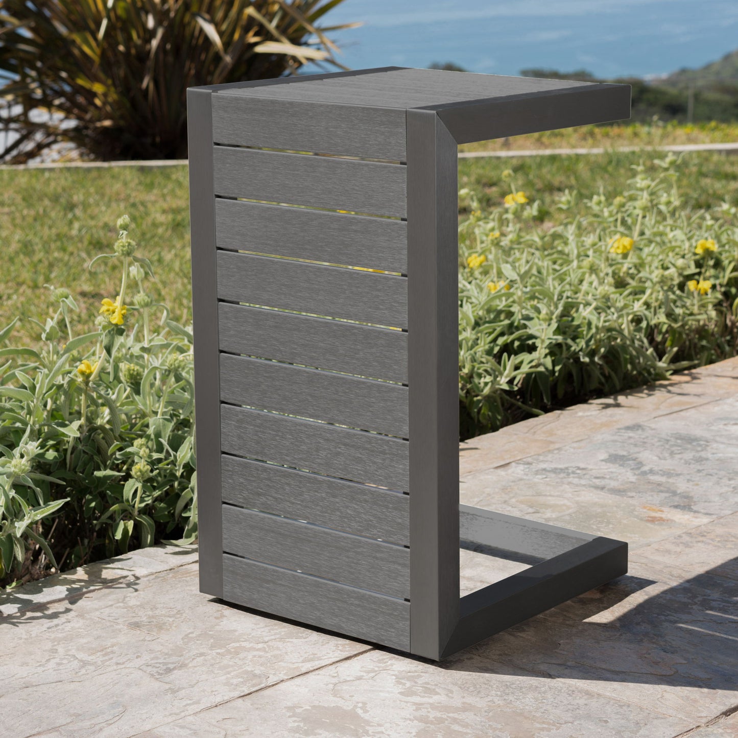 The Aluminum Outdoor C-Shaped Side Table for Patio, featuring horizontal slats and a modern gray finish, sits on a stone patio beside green plants, yellow flowers, grass, and an ocean view in the background.