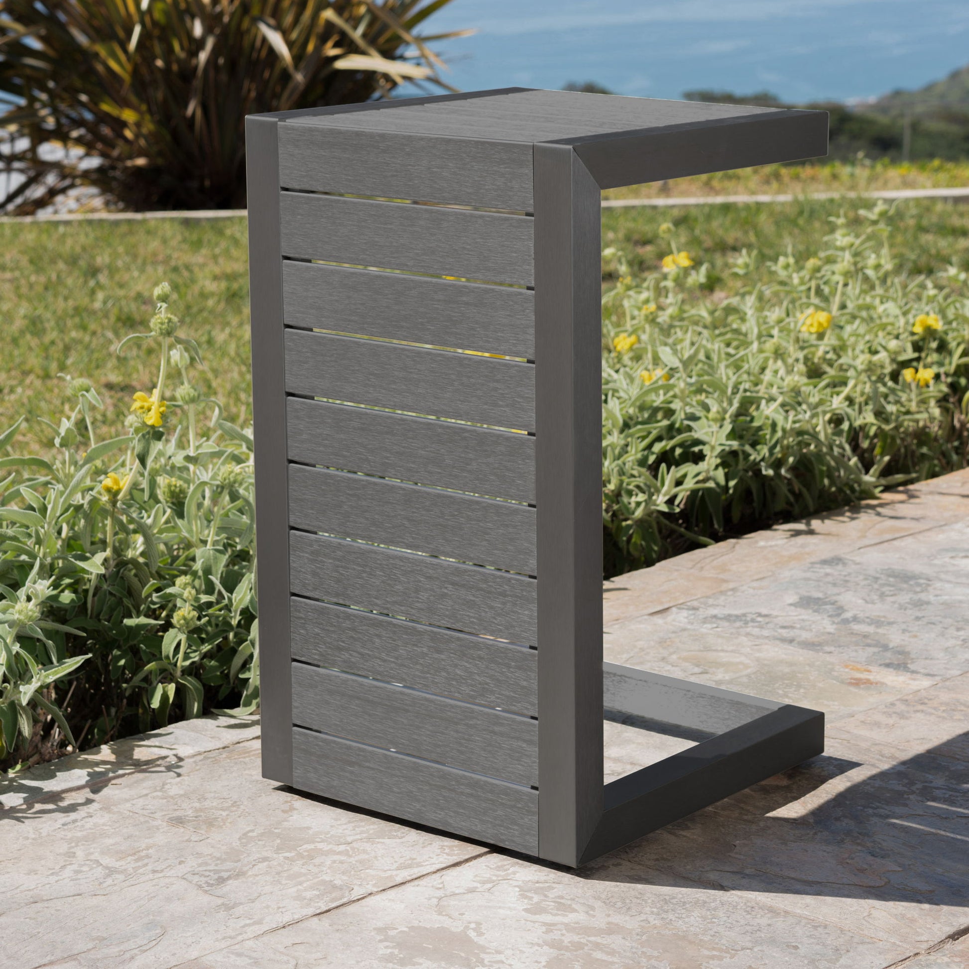 The Aluminum Outdoor C-Shaped Side Table for Patio, featuring horizontal slats and a modern gray finish, sits on a stone patio beside green plants, yellow flowers, grass, and an ocean view in the background.
